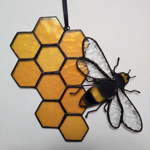 Amber stained glass 10 piece honeycomb with an irregular border .  To the one side a detailed bumble bee with wire twisted legs and black crystal eyes  made of stained glass in the colours of amber and black is overlaid and tack soldered to the right side of the honeycomb. The bees wings are open giving the appearance that it is flying away. The solder lines of both the  honeycomb and bee have had a black patina applied to them creating a richness and depth to the over piece.