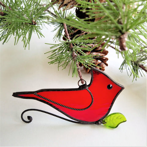 Stained glass red cardinal ornament, made from a slightly textured red art glass, with a jet black rhinestone eye and twisted wire wing detail. The wire and copper foil has a black patina. The cardinal is sitting on a curved wire branch with a green glass leaf attached .  The cardinal is displayed on a pine branch with pine cones attached.